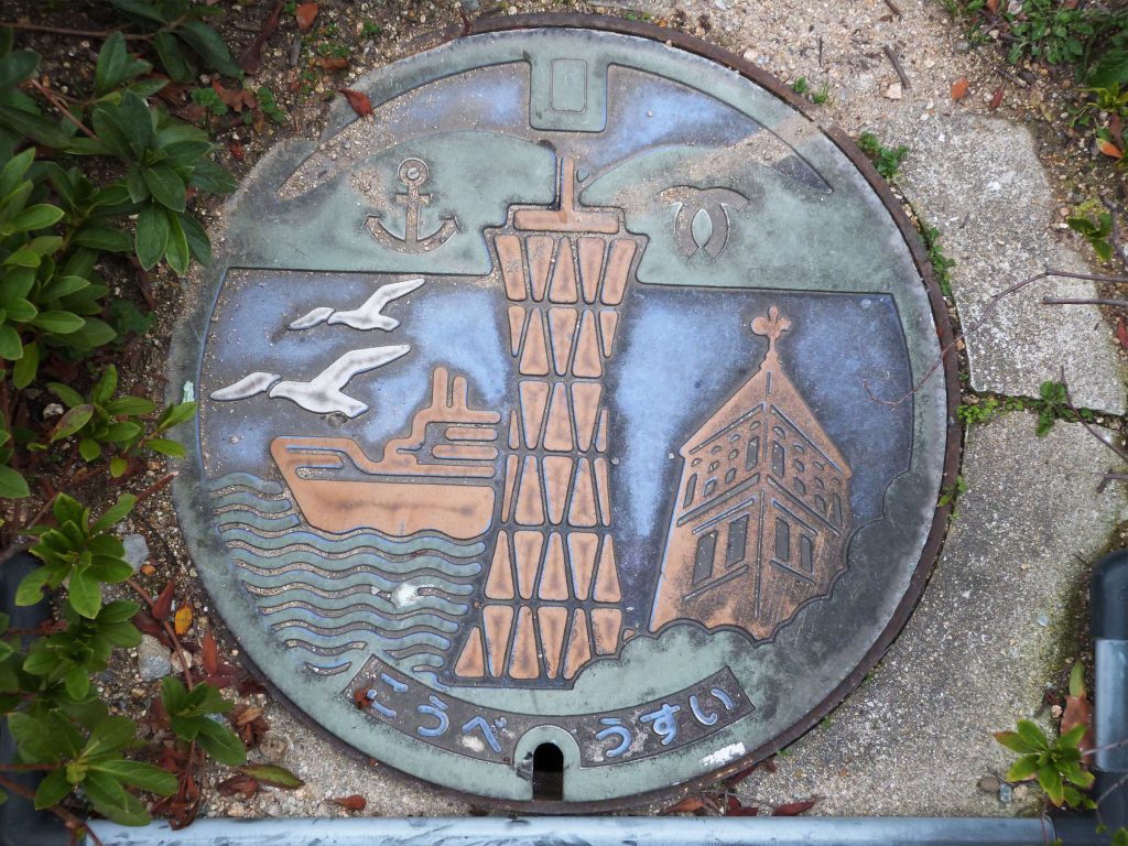 Couvercle de regard de chaussée de Kobe ; on où voit les collines de la ville et ses emblèmes, un paquebot, la tour du port et la girouette